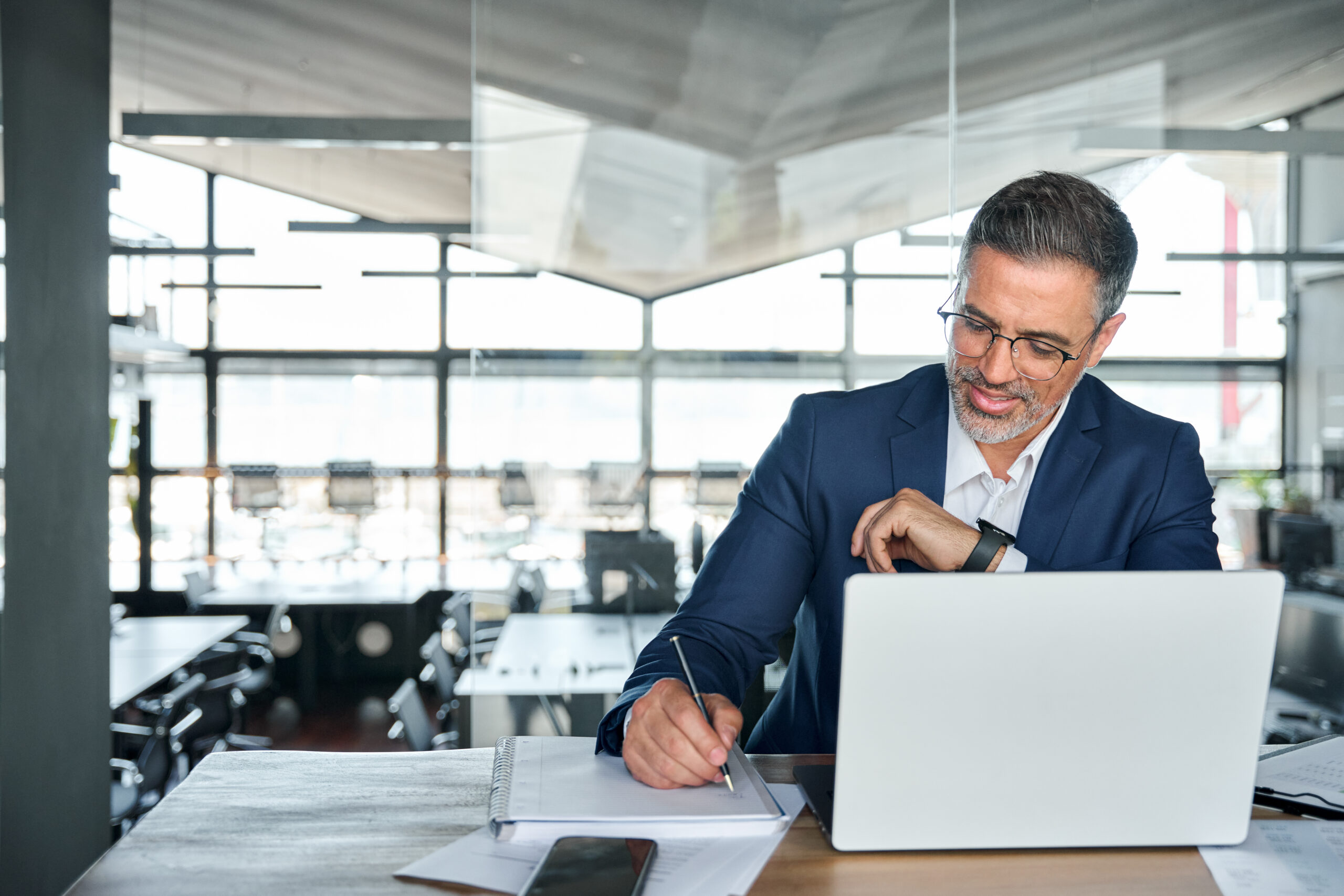 Middle aged smiling professional business man global company executive ceo manager or alawyer wearing suit sitting at desk in modern office working on laptop computer and writing notes, copy space.