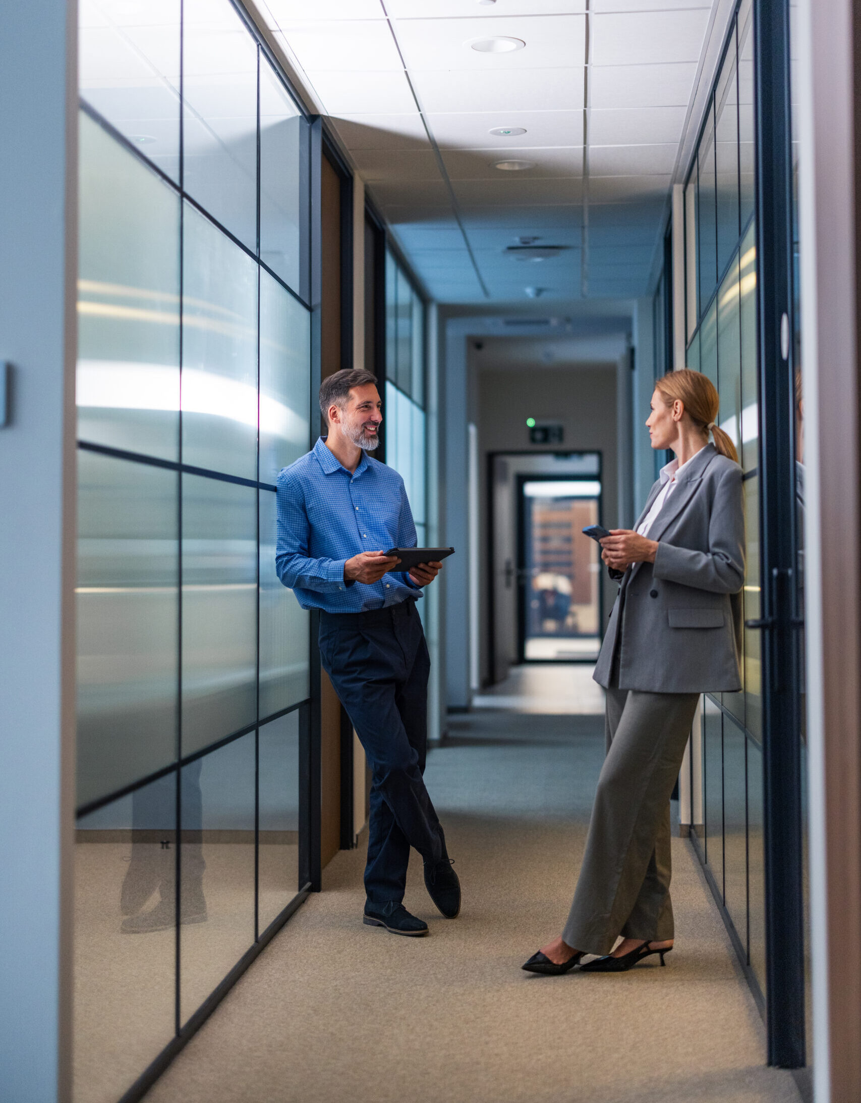 Two professionals chat in a glass-walled office corridor, using tablets during a casual work break.